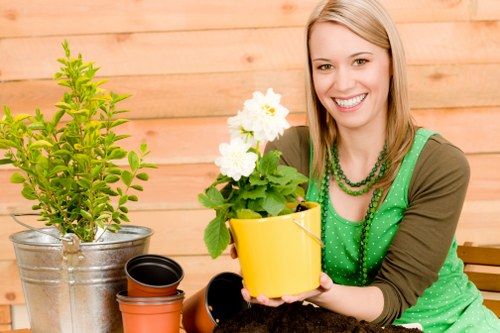 A woman with blonde hair, dressed in a green polka dot top and brown cardigan, sits outdoors in a garden setting holding a yellow flower pot containing a healthy, blooming white flower. In front of her, there are various gardening pots, including a silver pot with a small, lush green shrub and some empty terracotta pots. The background features a wooden fence, suggesting a neatly maintained backyard garden. The scene appears to be under natural daylight, with soft shadowing indicating a mild weather condition, suitable for outdoor gardening activities. This image illustrates a gardening enthusiast or professional engaging with plant care, emphasizing the importance of healthy potted plants and garden maintenance, aligning with services offered by Hedge Trimming Twickenham in the Twickenham area, post code TW1 or TW2, and demonstrates the care and attention involved in outdoor gardening and planting tasks.