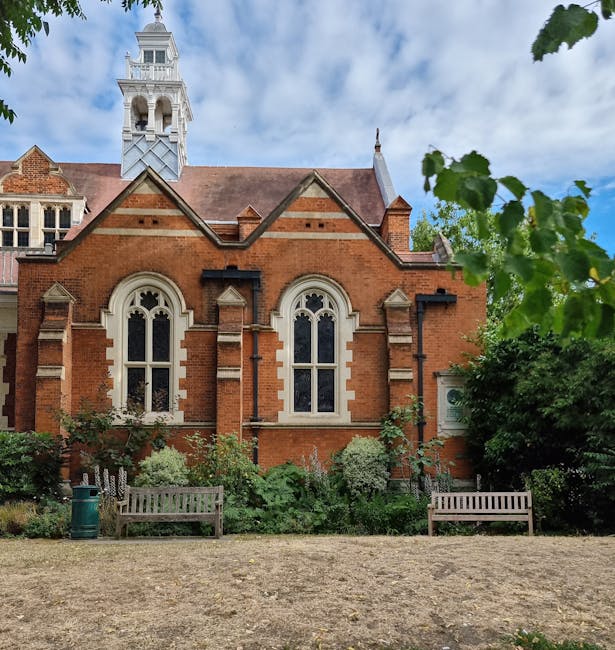 A historic red-brick church with pointed gothic windows framed by beige stone trims, set against a partly cloudy sky. The building features a decorative brick pattern and a small tower with a clock an